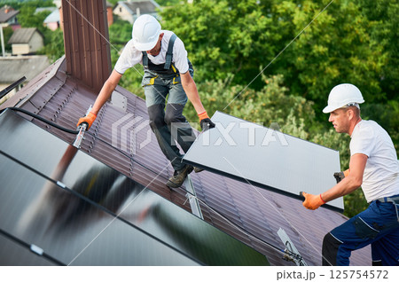 Workers building solar panel system on roof of house. Two men installers in helmets carrying photovoltaic solar module outdoors. Alternative, green and renewable energy generation concept. 125754572