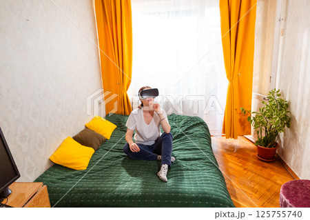 Female sitting on bed experiencing photorealistic digital landscapes through advanced VR technology. Wide shot, top view 125755740