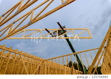 Workers use telehandler lift to position wooden truss in building under construction as clouds gather above. 125756995