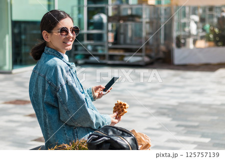 Carefree woman in her 20s snacks on a waffle while navigating vibrant city streets, merging youth travel, foodie moments, and weekend leisure Carefree woman in her 20s snacks on a waffle while navigating vibrant city streets, merging youth travel, foodie moments, and weekend leisure 125757139