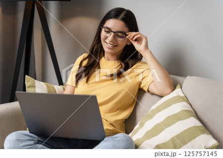 Woman sits comfortably on the couch in glasses, focused on her laptop as she works remotely, checks online orders, and scrolls through weekend deals Woman sits comfortably on the couch in glasses, focused on her laptop as she works remotely, checks online orders, and scrolls through weekend deals 125757145