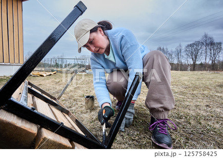 Treating underside of outdoor bench, attentive landscape renovator spreads black paint along metal supports, ensuring thorough coverage for weather resistance. 125758425