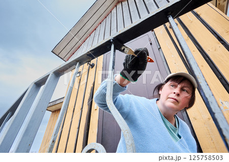 Applying protective paint to metal railings, woman carefully works on porch entrance of country house, focused on achieving smooth even coverage. Applying protective paint to metal railings, woman carefully works on porch entrance of country house, focused on achieving smooth even coverage. 125758503
