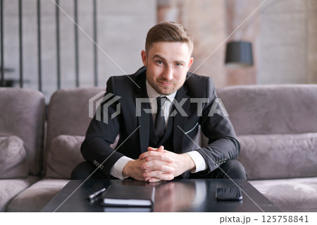 A man in an elegant black suit is sitting at a table with his arms folded and looking at the camera. The modern interior and soft lighting create an atmosphere of professionalism and confidence A man in an elegant black suit is sitting at a table with his arms folded and looking at the camera. The modern interior and soft lighting create an atmosphere of professionalism and confidence 125758841