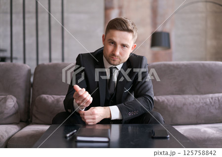 A man in a business suit is sitting at a table, pointing and looking away. The modern interior and soft lighting create a professional atmosphere. A man in a business suit is sitting at a table, pointing and looking away. The modern interior and soft lighting create a professional atmosphere. 125758842