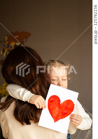Little daughter with golden hair presents hand-drawn card to her smiling mother. Genuine moment of family bonding and innocent affection. 125762156