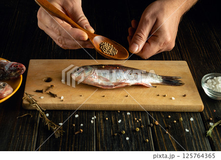 Hands are seasoning raw fish with mustard seeds, surrounded by salt and spices on a cutting board in a rustic kitchen. Fresh ingredients enhance the culinary experience 125764365
