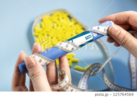 Female hands holding Ozempic syringe pen and measuring tape with a bunch of pills in the background 125764569