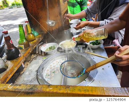 A street food vendor prepares mie ayam or Indonesian chicken noodle soup with meatballs, fresh greens, and broth, adding oil and ladling hot soup into bowls at a rustic outdoor stall. 125764904