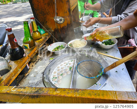 A street food vendor prepares mie ayam or Indonesian chicken noodle soup with meatballs, fresh greens, and broth, adding oil and ladling hot soup into bowls at a rustic outdoor stall. 125764905