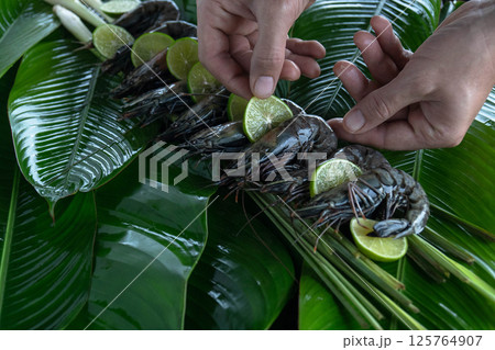 Hands skillfully place fresh tiger shrimp atop vibrant banana leaves, garnished with lime slices, creating an inviting display for a tropical feast. Hands skillfully place fresh tiger shrimp atop vibrant banana leaves, garnished with lime slices, creating an inviting display for a tropical feast. 125764907