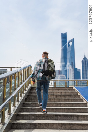 A young man wearing a plaid shirt, jeans, and sneakers walks up stairs with a backpack in Shanghai's Pudong, modern skyscrapers towering behind him A young man wearing a plaid shirt, jeans, and sneakers walks up stairs with a backpack in Shanghai's Pudong, modern skyscrapers towering behind him 125767604