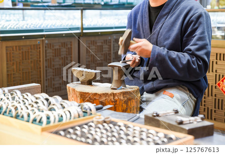 A craftsman in traditional blue jacket hand-forges jewelry rings on an anvil at Shanghai's Old Street Yunnan, with finished metal rings displayed in wooden trays at his open-air workshop A craftsman in traditional blue jacket hand-forges jewelry rings on an anvil at Shanghai's Old Street Yunnan, with finished metal rings displayed in wooden trays at his open-air workshop 125767613