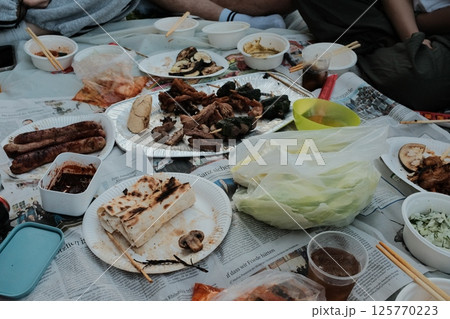 grilled meat and vegetables on barbecue grill and shared picnic food on blanket during outdoor gathering in park. High quality photo 125770223