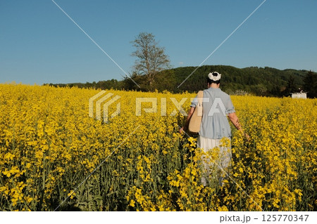 woman in white summer dress and straw hat walking through yellow rapeseed flower field under clear blue sky in countryside. High quality photo 125770347