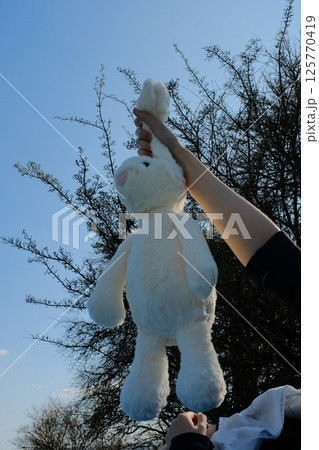 cute white plush rabbit toy held in hand against blue sky background in spring outdoor environment. High quality photo 125770419