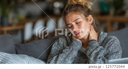 Tired young Caucasian woman sitting on sofa at home, holding her neck in pain. She appears stressed and fatigued, massaging her neck due to muscle tension. Tired young Caucasian woman sitting on sofa at home, holding her neck in pain. She appears stressed and fatigued, massaging her neck due to muscle tension. 125772044