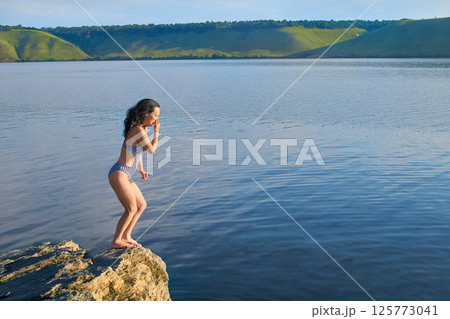 A young girl in swimsuit prepares to jump from cliff into the water of bay 125773041