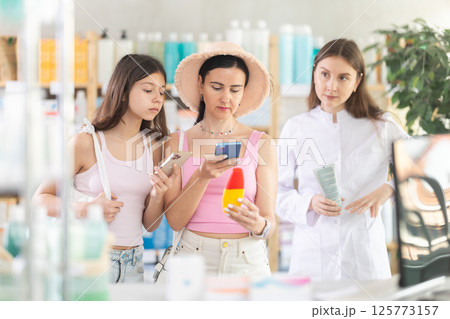Mother and daughter selecting sunscreen cream or sun protection product and scan QR code in drugstore, woman pharmacist consulting customers 125773157