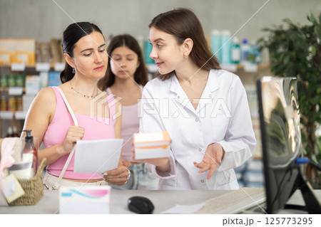 Young professional female parmacist working computer and helping mother and daughter clients with prescription in hand look for medicine in drugstore Young professional female parmacist working computer and helping mother and daughter clients with prescription in hand look for medicine in drugstore 125773295