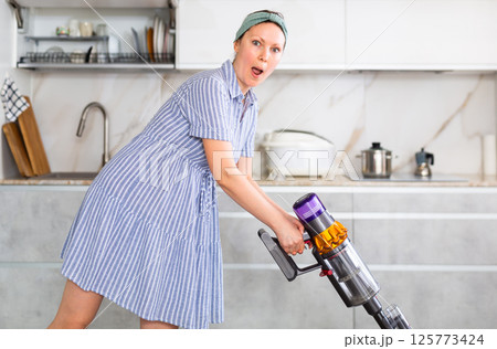 Surprised housewife with vacuum cleaner cleans floor covering near dining table Surprised housewife with vacuum cleaner cleans floor covering near dining table 125773424