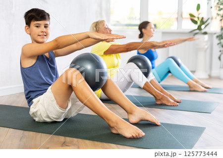 Preteen boy practicing fitness with softball between legs in light training room 125773444