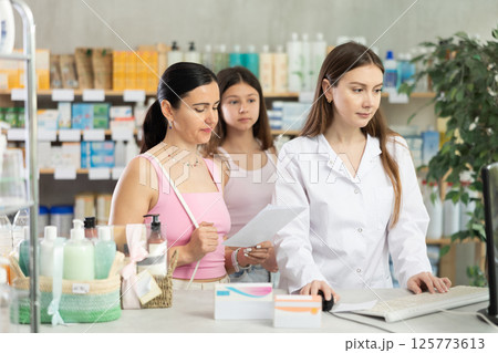 Young professional female parmacist working computer and helping mother and daughter clients with prescription in hand look for medicine in drugstore Young professional female parmacist working computer and helping mother and daughter clients with prescription in hand look for medicine in drugstore 125773613