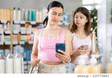 Mom and daughter scrolling phone while looking for something in drugstore 125773661