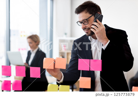 Young male professional wearing formal suit organizing tasks using sticky notes. Transparent glass board displays structured planning. Female colleague working with laptop in office environment. 125773879
