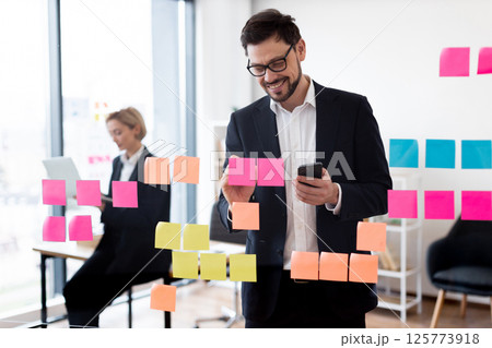 Business individuals, man and woman, collaborating on project using colorful notes on glass board. Scene depicts teamwork and planning in professional setting with modern office environment Business individuals, man and woman, collaborating on project using colorful notes on glass board. Scene depicts teamwork and planning in professional setting with modern office environment 125773918