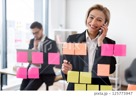 Businesswoman discussing strategy on phone near glass with sticky notes, colleague working background Businesswoman discussing strategy on phone near glass with sticky notes, colleague working background 125773947