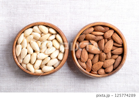 Almonds with skin and skinless almonds in wooden bowls on linen. Fruits of Prunus amygdalus, syn. Prunus dulcis, with seed coats and blanched, ready-to-eat as snack or as ingredient for baking. Photo 125774289