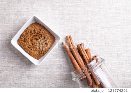 Cassia cinnamon powder in a white bowl, with cinnamon sticks sticking out of an open screw-top jar, on linen fabric, with empty space for text. Chinese cinnamon, Cinnamomum cassia, used as a spice. 125774291