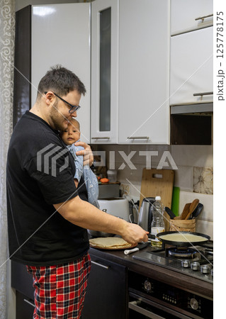 Father Cooking Pancakes While Holding Baby Father Cooking Pancakes While Holding Baby 125778175