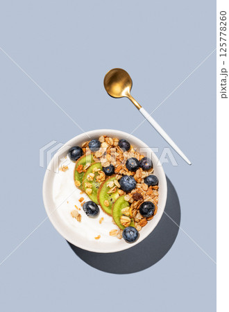 Natural yogurt with granola, kiwi and blueberries in a bowl on a blue background with spoon. 125778260