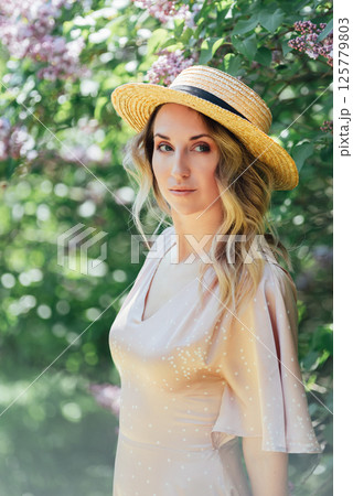 Young Woman In Summer Hat Enjoying The Fragrance Of Lilac Blooms On Sunny Day. lady in a straw hat and dress smelling lilac flowers in spring sunlit garden. 125779803