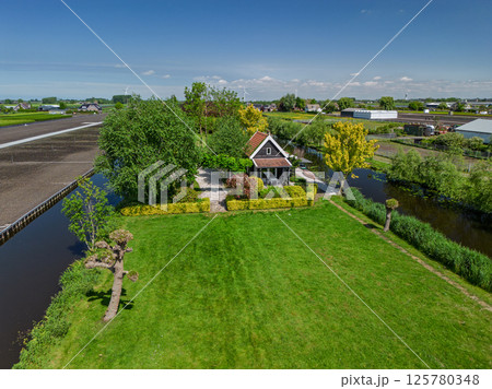 Charming countryside house surrounded by lush green gardens, trees, and canals under a clear blue sky, located in a peaceful rural landscape with farms and greenhouses in the distance. 125780348