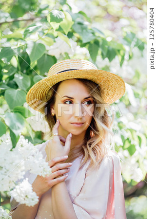 Young Woman In Summer Hat Enjoying The Fragrance Of Lilac Blooms On Sunny Day. lady in a straw hat and dress smelling lilac flowers in spring sunlit garden. Young Woman In Summer Hat Enjoying The Fragrance Of Lilac Blooms On Sunny Day. lady in a straw hat and dress smelling lilac flowers in spring sunlit garden. 125780404