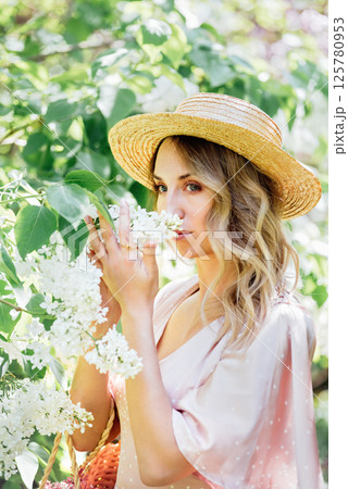 Woman in straw hat and pink dress gently smelling white lilac flowers in lush green garden on a sunny spring day Woman in straw hat and pink dress gently smelling white lilac flowers in lush green garden on a sunny spring day 125780953