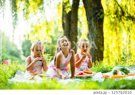 The sisters are enjoying their time together, sitting on a soft picnic blanket. The twins, dressed alike, laugh as they eat their watermelon, while their older sister playfully joins in. The sisters are enjoying their time together, sitting on a soft picnic blanket. The twins, dressed alike, laugh as they eat their watermelon, while their older sister playfully joins in. 125782010