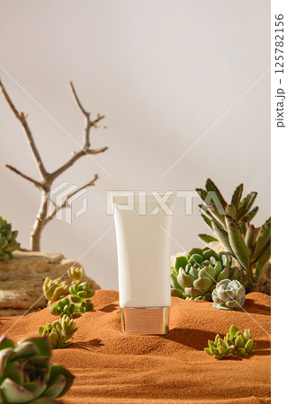 Unbranded white cosmetic tube with shiny gold plastic cap. Some succulents and cactuses are placed around the sandy beach. Rocks and dry branches blurred in the background. White background. 125782156