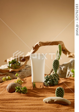 Mockup of white cosmetic tube with gold cap, standing on the center of the dry sand. Natural light shines in, shadows of objects fall on the sand. Spiny cactus grows around, interspersed with pebbles 125782163