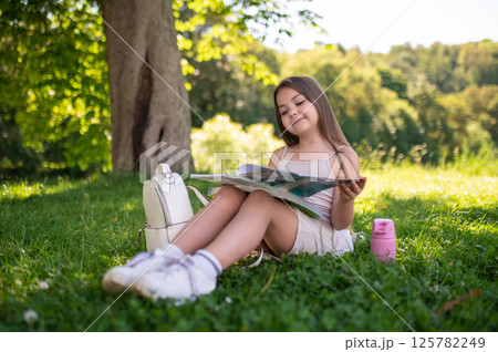 Cheerful kid sitting on grass with book in her hands reading fun story Cheerful kid sitting on grass with book in her hands reading fun story 125782249