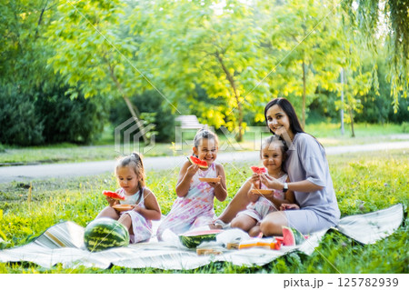 The three sisters sit with their mother on a picnic blanket. The twins giggle as they bite into juicy watermelon. 125782939
