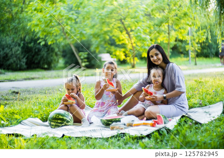 The three sisters sit close with their mother, sharing a big watermelon slice. The twins giggle, their faces sticky with juice. 125782945