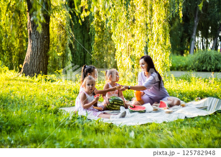 The girls and their mother enjoy a sunny picnic. The twins take big bites of watermelon, laughing. 125782948