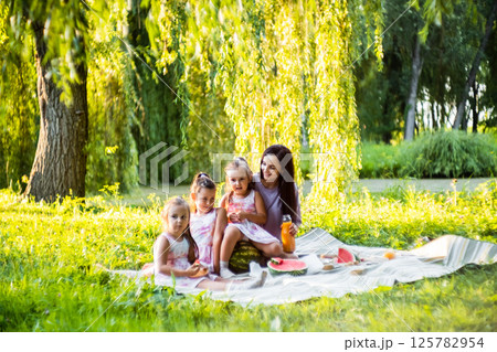 The three sisters sit around their mother, enjoying the picnic. The twins smile as they take bites of watermelon.  125782954