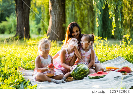The girls in matching dresses sit on a blanket with their mom. The twins eat watermelon, giggling at the mess. 125782968