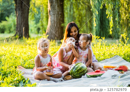 The girls in matching dresses sit on a blanket with their mom. The twins eat watermelon, giggling at the mess. 125782970