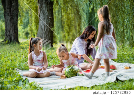 The girls laugh and eat watermelon in the park. The twins try to take the biggest bites. 125782980